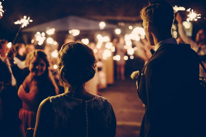 A bride and groom walk out of their reception, where the crowd all hold sparklers