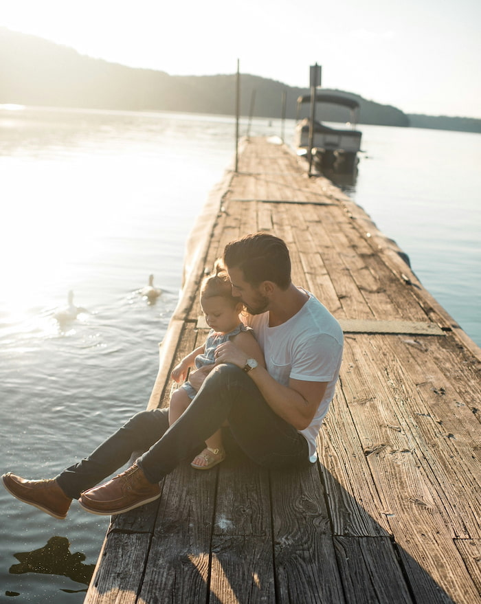 A man holds his young daughter, both sat on a pier while ducks swim in the water nearby