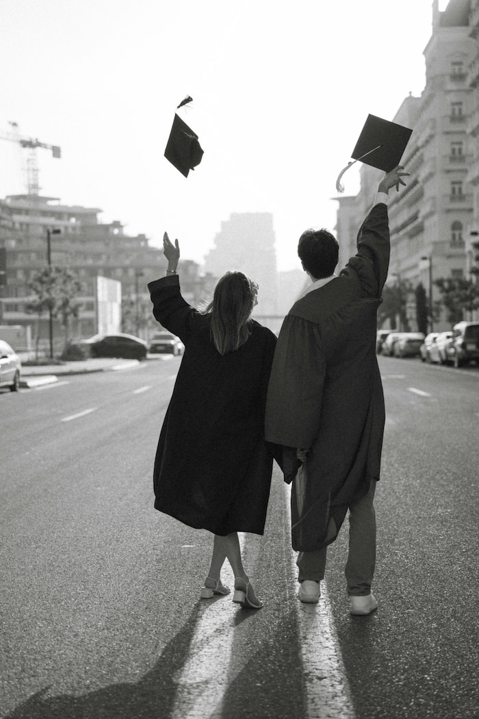 A young couple walk down the street in graduation gowns, throwing their caps in the air