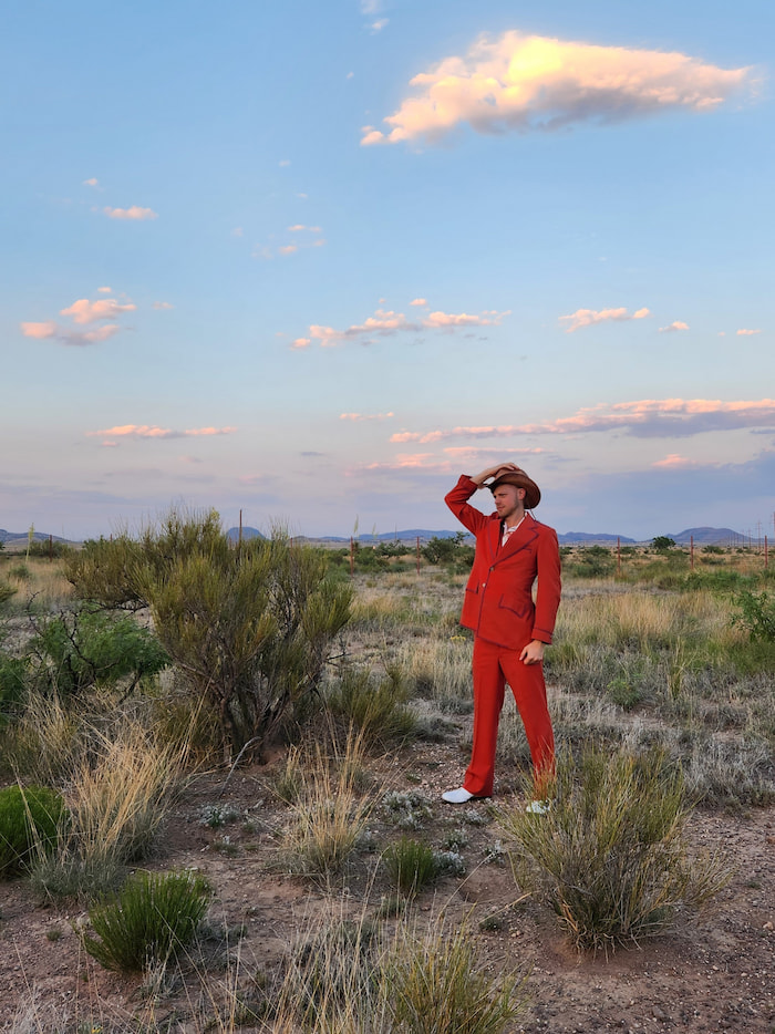 A man in a vintage red suit and cowboy hat standing in a desert with mountains in the background